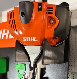 Wzmocniony pomarańczowy uchwyt ścienny z druku 3D do profesjonalnej kosy spalinowej STIHL FS 460 zamontowany na ścianie.
